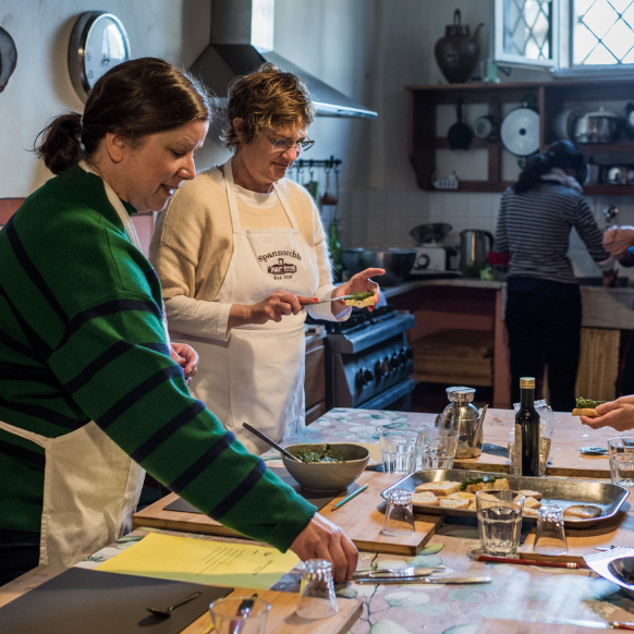 A photo of two people wearing aprons and standing at a big table filled with various kitchen and cookware and food items. Behind them is a stove and the back of another person standing at a sink, with some shelving filled with more kitchen items on the wall.