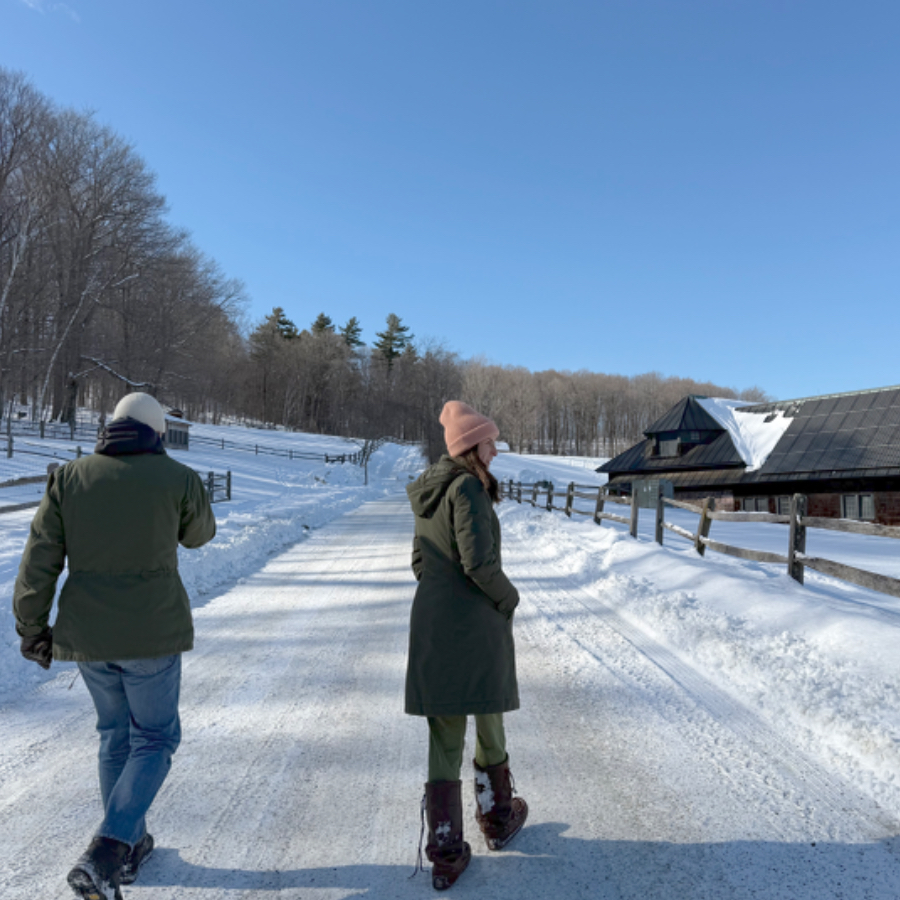 Two people walking along a snowy road with fields on both sides and an old farm building on the right. Bare trees and clear blue sky are in the background.